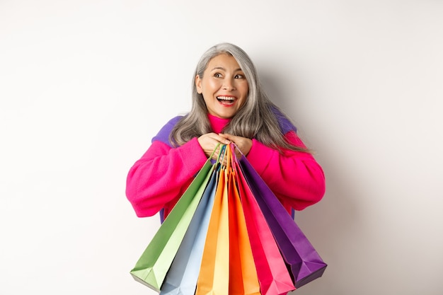 買い物中毒のファッショナブルなシニアアジアの女性、買い物袋を抱き締めて、うれしそうな笑顔、割引で購入、白い背景の上に立って