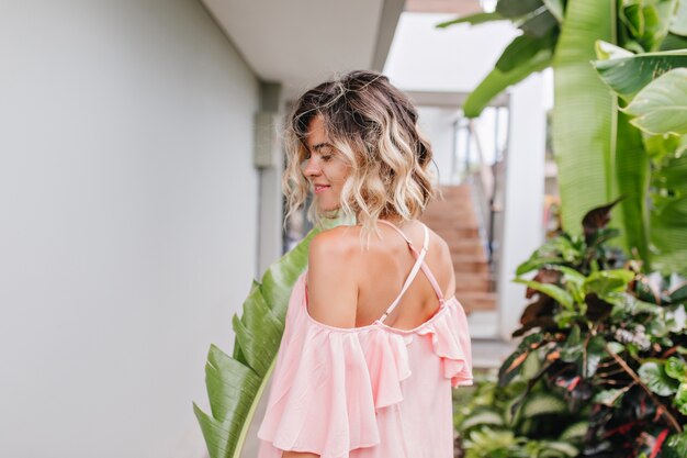 Fashionable short-haired girl walking by green bushes. Outdoor photo from back of stylish blonde woman wears pink clothes.