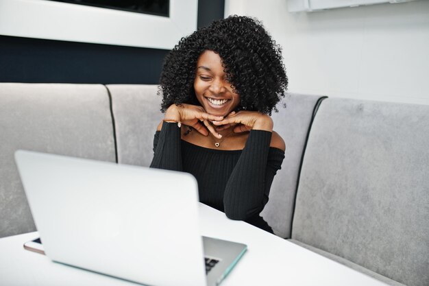 Модная улыбающаяся молодая красивая афроамериканская деловая женщина с афро-прической в элегантном черном сидит и работает за ноутбуком