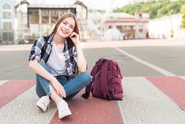 Fashionable smiling woman sitting on road with her backpack