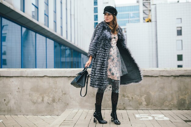 Модная улыбающаяся женщина гуляет по городу в теплой шубе, зимний сезон, холодная погода, в черной кепке, платье, сапогах, держит кожаную сумку, тренд уличной моды