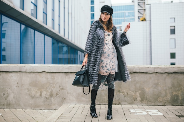 Donna sorridente alla moda che cammina in città in pelliccia calda, stagione invernale, clima freddo, berretto nero, vestito, stivali, borsa in pelle, tendenza della moda di strada