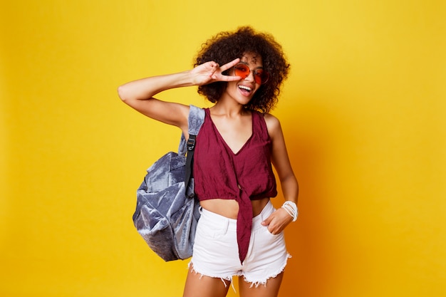 Immagine alla moda dello studio della femmina nera graziosa sexy con i capelli di afro. vestito estivo, zaino e occhiali da sole. donna adorabile che balla su sfondo giallo.