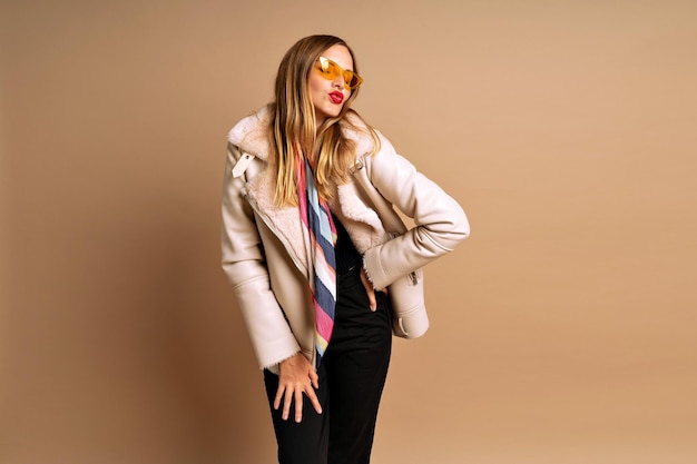 Free photo fashionable studio portrait of beautiful blonde woman wearing fur trendy jacket and colorful scarf, beige background, autumn winter fall season