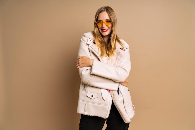 Модный студийный портрет красивой блондинки в меховой модной куртке и красочном шарфе на бежевом фоне осенне-зимний осенний сезон