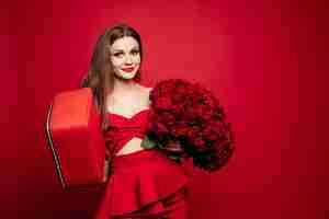 Free photo fashionable studio portrait of stylish young woman with long brown hair in expensive red suit with leather red bag and bunch of red roses she is smiling at camera red lips red background