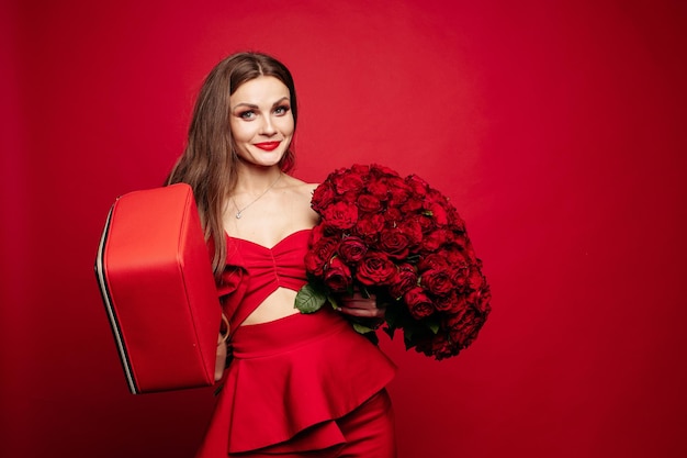 Fashionable studio portrait of stylish young woman with long brown hair in expensive red suit with leather red bag and bunch of red roses She is smiling at camera Red lips Red background