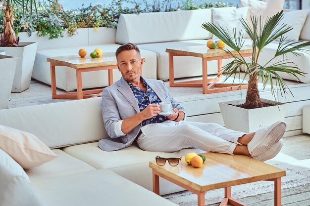 Fashionable successful man with stylish hair dressed in modern elegant clothes holds cup of coffee while sitting on a sofa at outdoor cafe against the background of the city wharf.