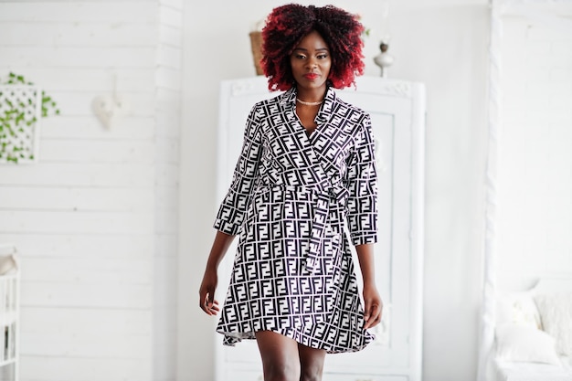 Free photo fashionable tall african american model woman with red afro hair in dress posed at white room against vintage wardrobe