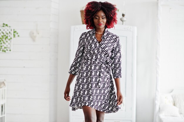 Fashionable tall african american model woman with red afro hair in dress posed at white room against vintage wardrobe
