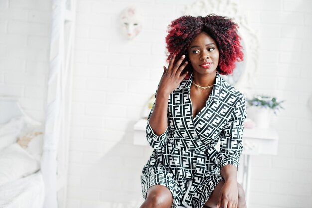 Fashionable tall african american model woman with red afro hair in dress posed at white room sitting on chair against mirror