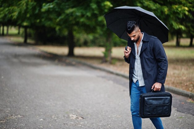 비오는 날에 포즈를 취한 우산과 가방 케이스가 있는 검은 코트를 입은 세련된 키 큰 아랍 수염 남자