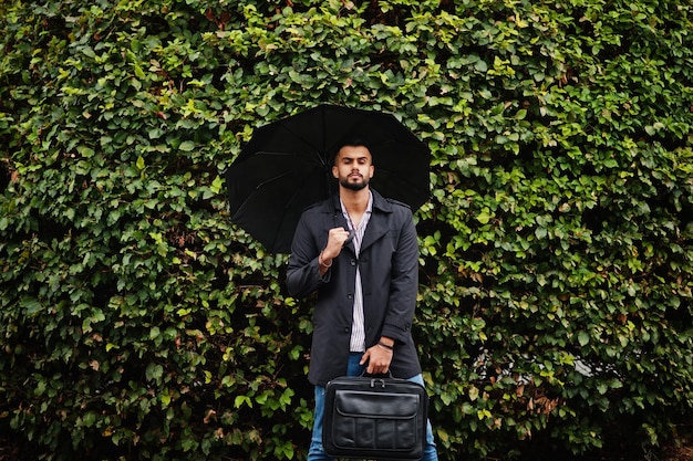 Foto gratuita l'uomo alla moda con barba araba alta indossa un cappotto nero con ombrello e custodia per borsa poste in una giornata di pioggia