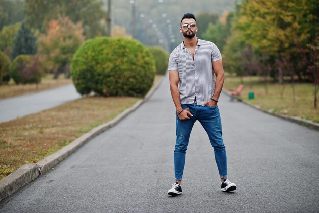 Модный высокий арабский бородатый мужчина в джинсах-рубашках и солнцезащитных очках позирует в парке и держит кошелек под рукой
