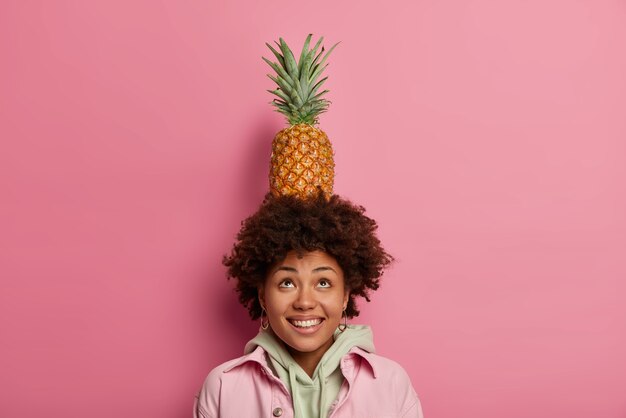 Fashionable teenager holds pineapple on head, makes trick with tropical fruit, looks above, smiles broadly, has white teeth, wears hoodie with jacket