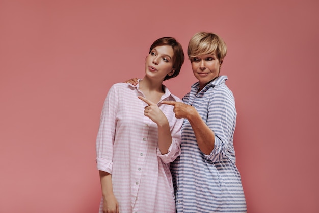 Foto gratuita alla moda due donne con acconciatura corta ed elegante in abiti freddi a strisce che mostrano il posto per il testo su sfondo rosa isolato.