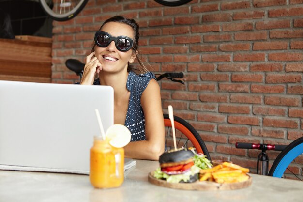 Fashionable video blogger in stylish sunglasses recording webcam video on laptop pc sitting against red brick wall of modern cafe. Happy woman with pretty smile surfing internet on notebook computer