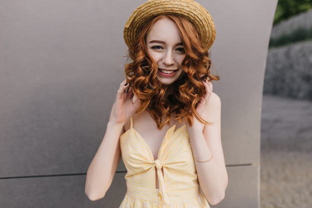 Fashionable white lady in cute hat posing with shy smile. Portrait of adorable curly woman with pale skin and ginger hair.