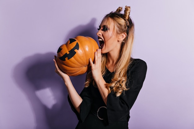 Fashionable witch screaming on purple wall. Magnificent vampire girl looking at pumpkin.