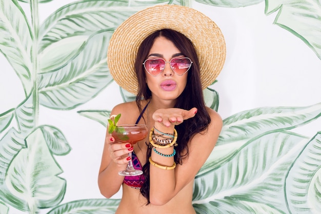 Fashionable woman in bright pink heart sunglasses drink fresh strawberry cocktail in beach bar.