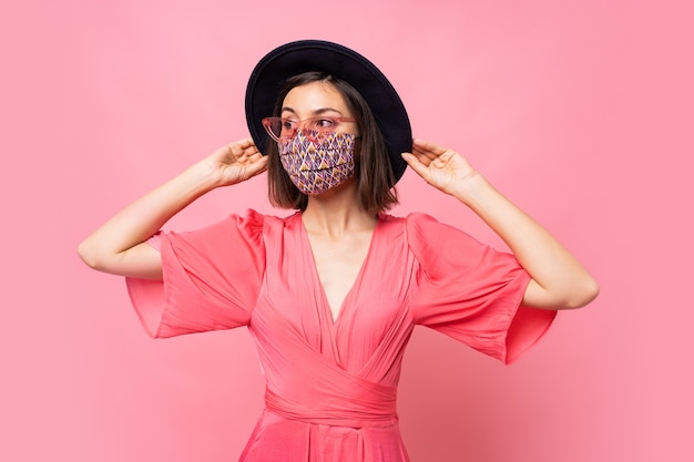 Donna alla moda vestita maschera protettiva elegante. indossare cappello nero e occhiali da sole. in posa sul muro rosa