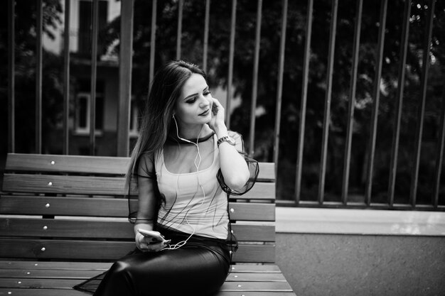 ファッショナブルな女性は、携帯電話とヘッドフォンで通りにポーズをとってベンチに座っている白いシャツ黒の透明な服の革のズボンを見てモバイルで音楽を聴くファッションの女の子の概念