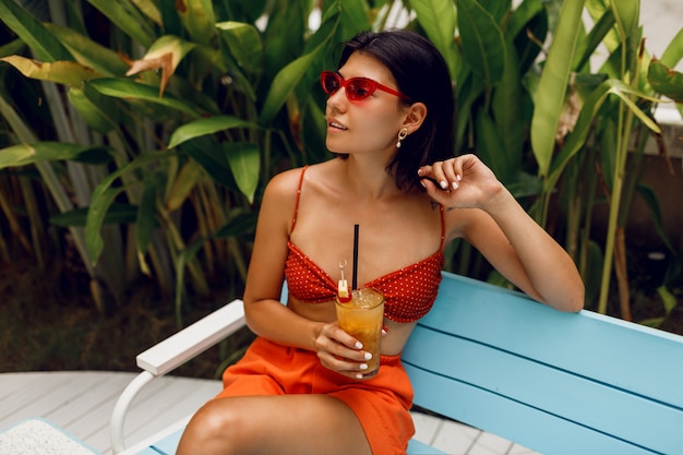 スタイリッシュな赤いトップとオレンジのショートパンツのトロピカルビーチクラブでリラックスしたレトロなサングラスでファッショナブルな女性。おいしいレモネードを飲みます。