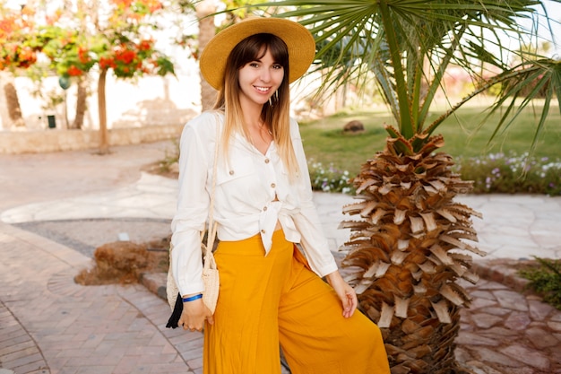 Fashionable woman standing on palms and blooming trees Wearing straw hat. Holidays and travel concept.