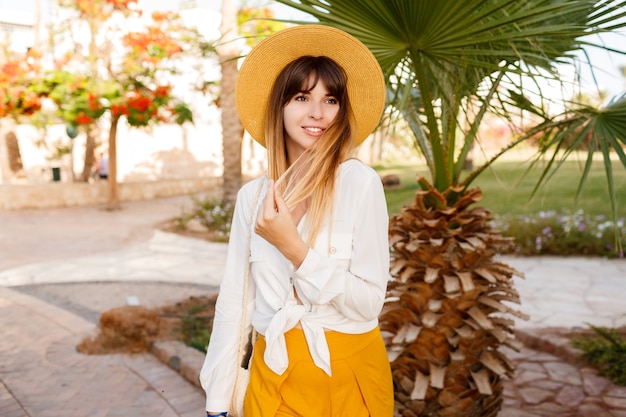 Fashionable woman standing on palms and blooming trees wearing straw hat.