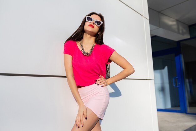 Модная женщина в летней одежде и солнцезащитных очках позирует над белой городской стеной