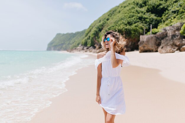熱帯の島で時間を過ごす白い服を着たファッショナブルな女性。リゾートで自然の景色を楽しむ魅力的な金髪の女性の屋外の肖像画。
