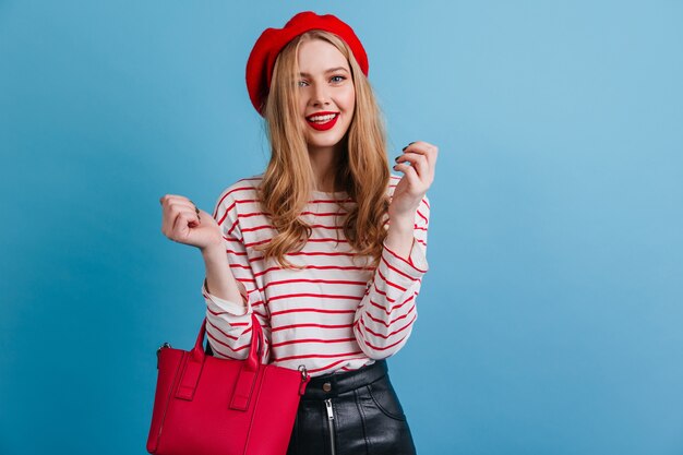 Fashionable woman with handbag standing on blue wall. Front view of playful blonde girl in french beret.