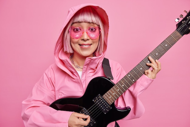 Fashionable woman with pink hair pretends performing on stage plays rock and roll music wears heart shaped sunglasses and anorak poses indoor against rosy wall. Talented female soloist