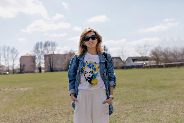 Free photo fashionable woman with short hairstyle is wearing sunglasses and white clothes and denim jacket is posing at camera on background of field and blue sky in sunlight