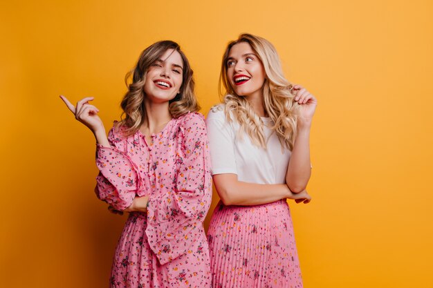 Fashionable woman with wavy hair spending time with sister. pleased female friends standing on yellow wall.