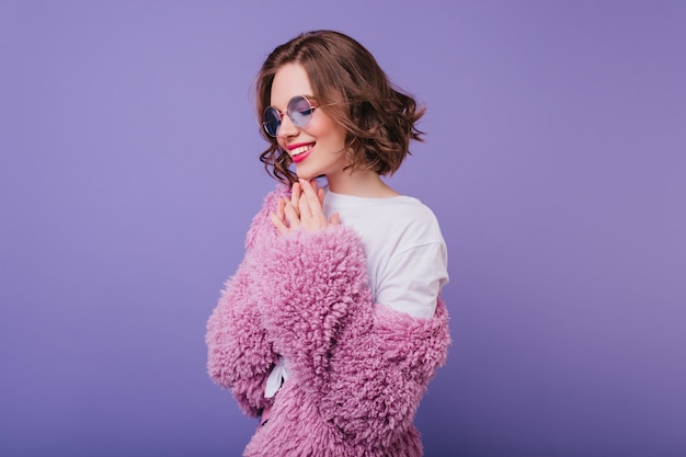 Fashionable woman with wavy hairstyle smiling with eyes closed. Indoor photo of enthusiastic female model in pink fur jacket.