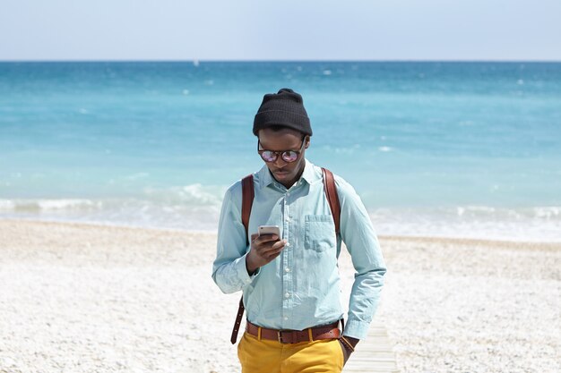 Fashionable young African American male tourist using mobile phone on desert beach, posting pictures of beautiful seascape around him via social media with azure ocean and blue sky in horizon
