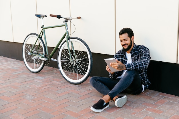 おしゃれな若いひげのある男は、デジタルタブレットを使用して、明るく笑って