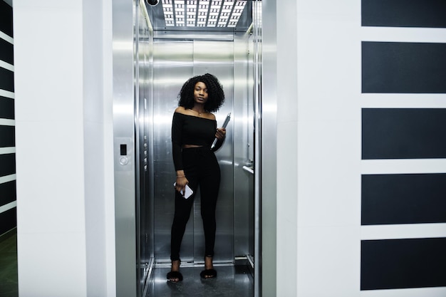 Fashionable young beautiful african american business woman with afro hairstyle wear in elegant black stand at elevator with phone and laptop at hands