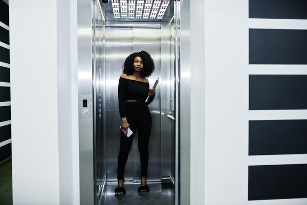 Fashionable young beautiful african american business woman with afro hairstyle wear in elegant black stand at elevator with phone and laptop at hands