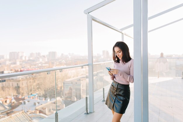 Fashionable young brunette woman in black skirt using phone on terrace on city view.
