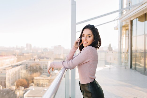 Fashionable young brunette woman speaking on phone on terrace on city view in sunny morning. Success, luxury lifestyle, chilling, relaxing, joyful businesswoman, smiling to side.