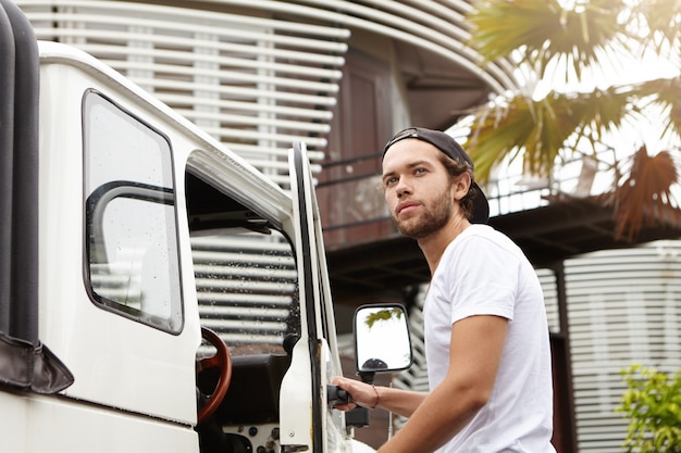 Fashionable young Caucasian male student wearing snapback opening door of his white four-wheel drive vehicle, looking away with smile