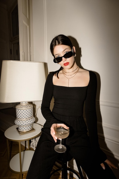 Fashionable young caucasian woman in black glasses and clothes looks at camera sitting indoors with glass of booze Stylish lady having a good time