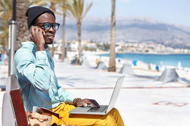 Fashionable young dark-skinned freelancer in hat and sunglasses having phone conversation on mobile while working remotely on laptop pc, sitting on bench in urban beach surroundings during holiday