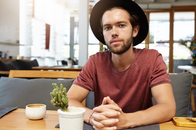 Fashionable young entrepreneur wearing t-shirt and trendy black hat sitting at wooden table with mug and cactus while having cappuccino at coffee shop alone, waiting for his partner for meeting