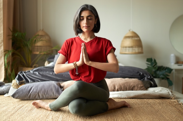 Fashionable young female with grayish hair sitting in gomukhasana or cow pose while practicing hatha yoga in bedroom after awakening, keeping eyes closed, pressing hands together in namaste