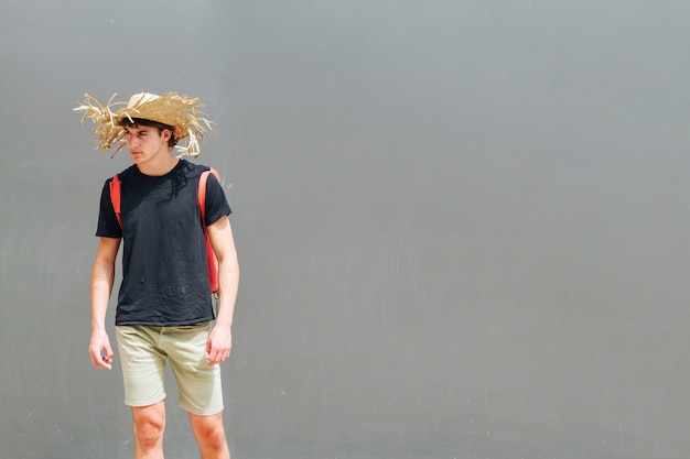 Foto gratuita giovane uomo bello alla moda con il cappello di paglia e lo zaino che stanno contro il fondo grigio