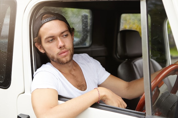 Fashionable young man wearing snapback backwards driving his sport utility vehicle and sticking his head and elbow out of open window, looking at road with concerned expression