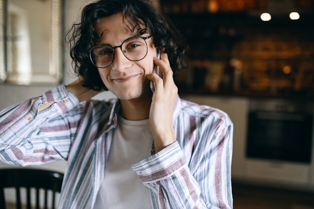 Fashionable young man with black curls smiling at camera while making phone call.